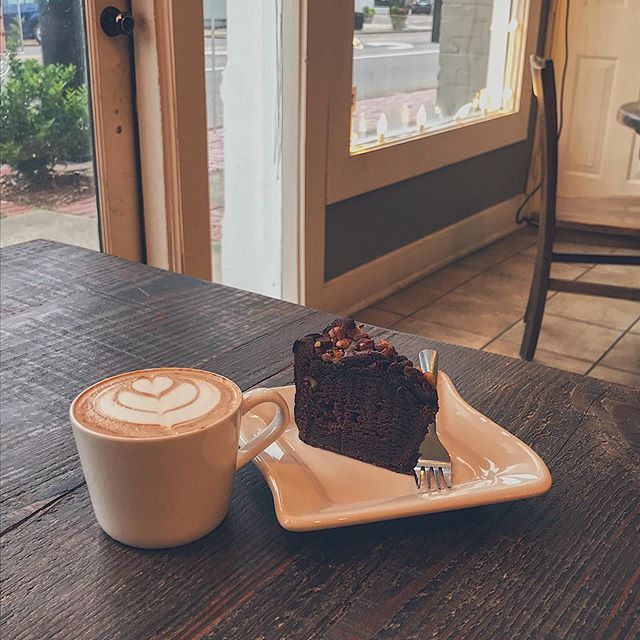 A cup of latte with heart-shaped foam art and a slice of chocolate cake on a plate sit on a dark wooden table.