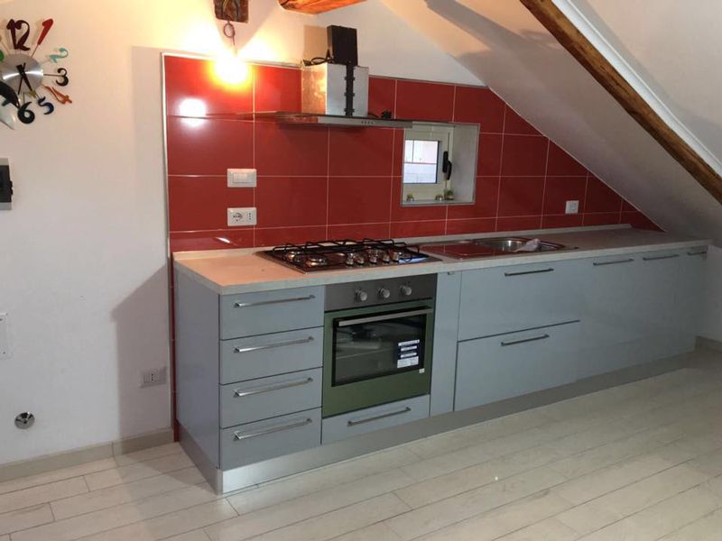 Cucina moderna e compatta con paraschizzi rosso, mobili grigi e soffitto inclinato.