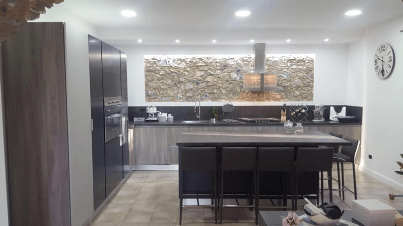 Cucina moderna con isola, sgabelli da bar scuri, paraschizzi in pietra e faretti a incasso a soffitto.