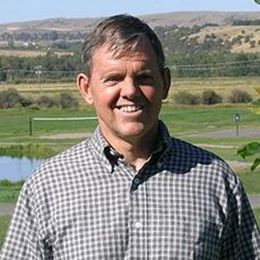 A man in a plaid shirt is smiling in front of a field.