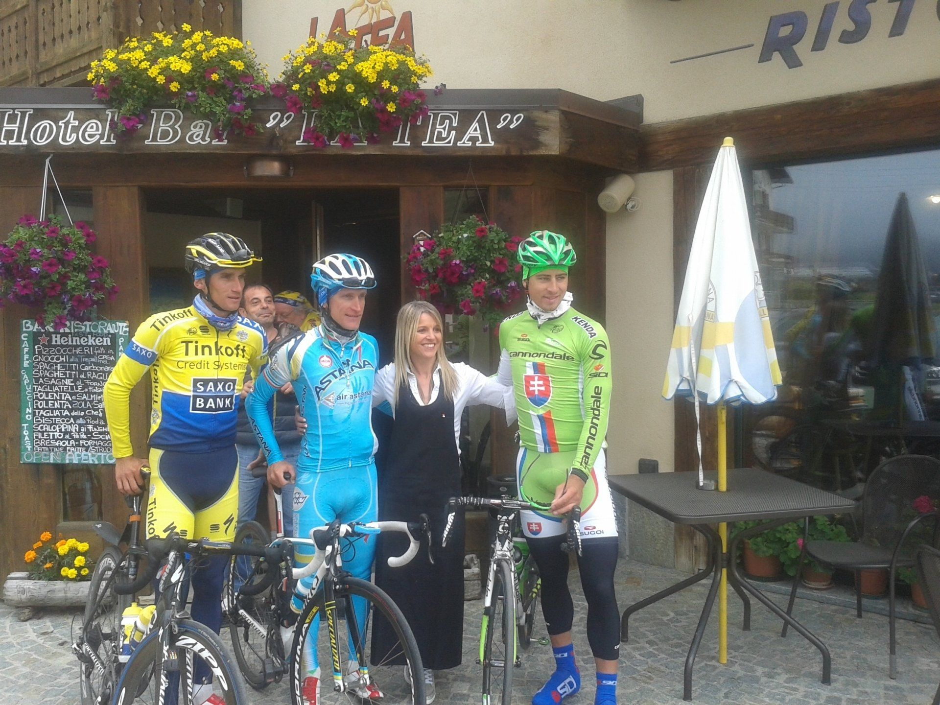 Ciclisti con maglie dai colori vivaci posano con una donna fuori da un ristorante, con le biciclette davanti.