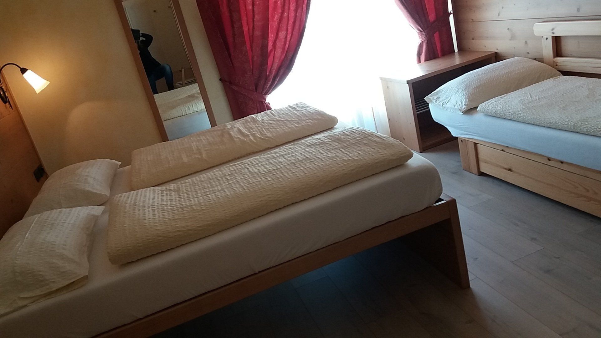 Due letti singoli in una camera da letto con tende rosse. Una lampada è appesa al muro.