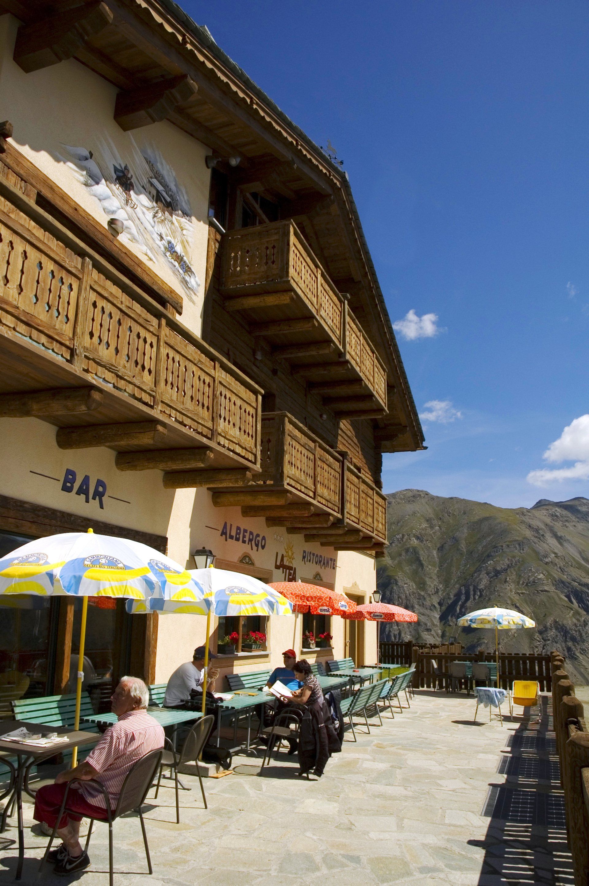 Persone sedute ai tavoli all'aperto, sotto gli ombrelloni, in un ristorante di montagna, in una giornata di sole.