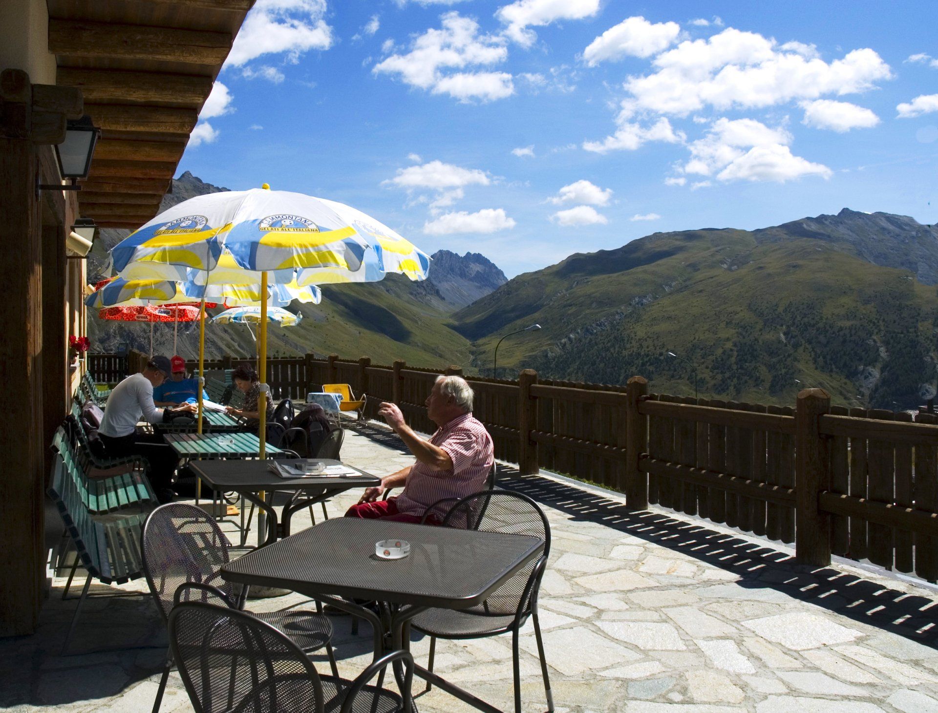 Persone sedute ai tavolini dei bar all'aperto con vista sulle montagne, sotto gli ombrelloni, in una giornata di sole.