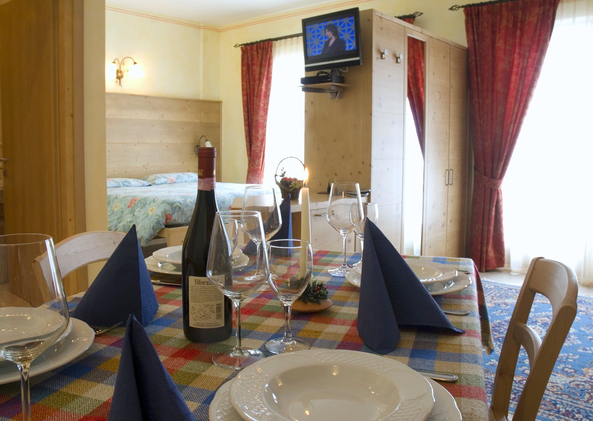 Tavolo da pranzo apparecchiato per un pasto, con bottiglia di vino, bicchieri e tovaglioli blu, in una stanza con un letto.