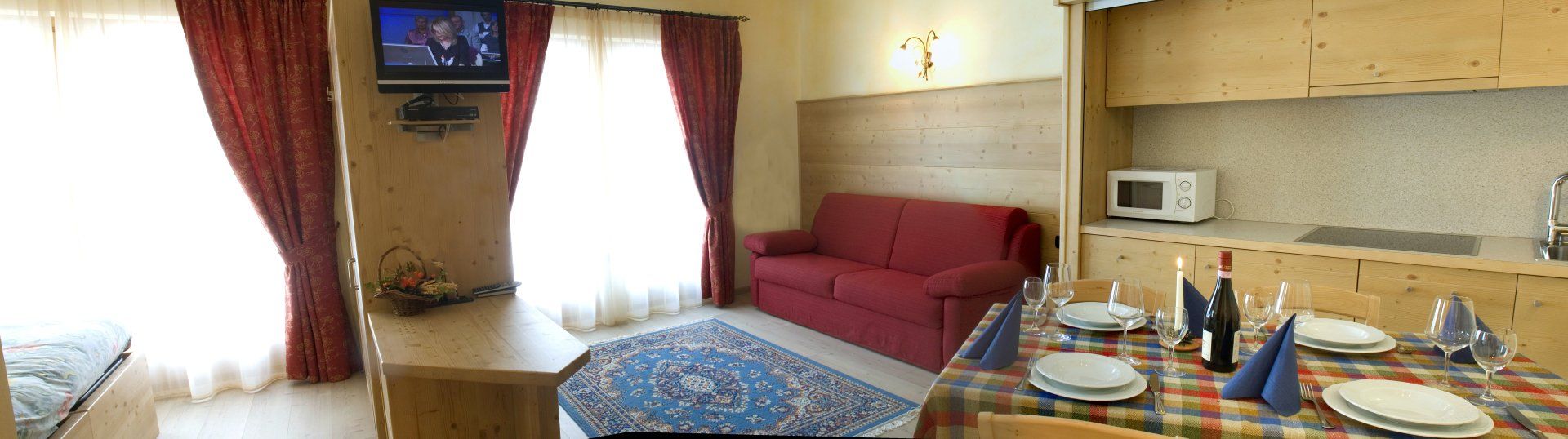 Zona giorno con divano rosso, TV, tavolo da pranzo e angolo cottura.