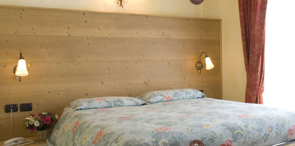 Camera da letto con testiera in legno, due lampade da comodino e copriletto a motivi floreali. Tenda rossa sulla destra.