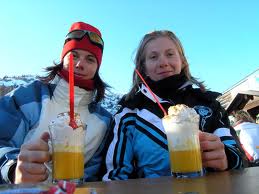 Due donne in abiti invernali sedute a un tavolo, con in mano dei drink gialli con panna montata, cannucce rosse e un cielo azzurro sullo sfondo.