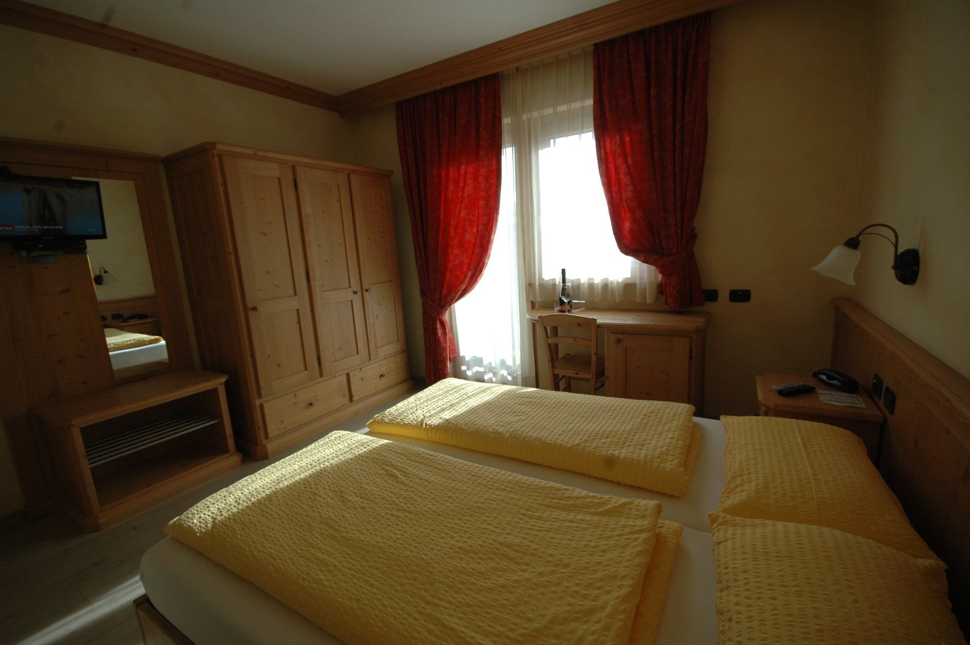 Camera da letto con due letti, mobili in legno, tende rosse e una piccola finestra.
