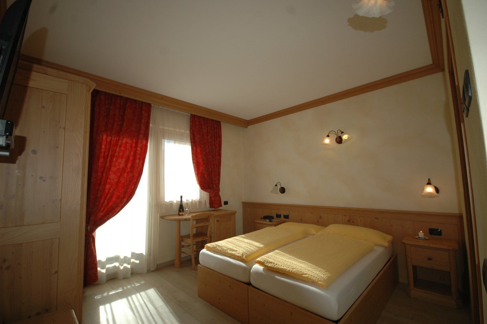 Camera da letto con due letti, tende rosse, tavolino vicino alla finestra, pareti chiare e mobili in legno.