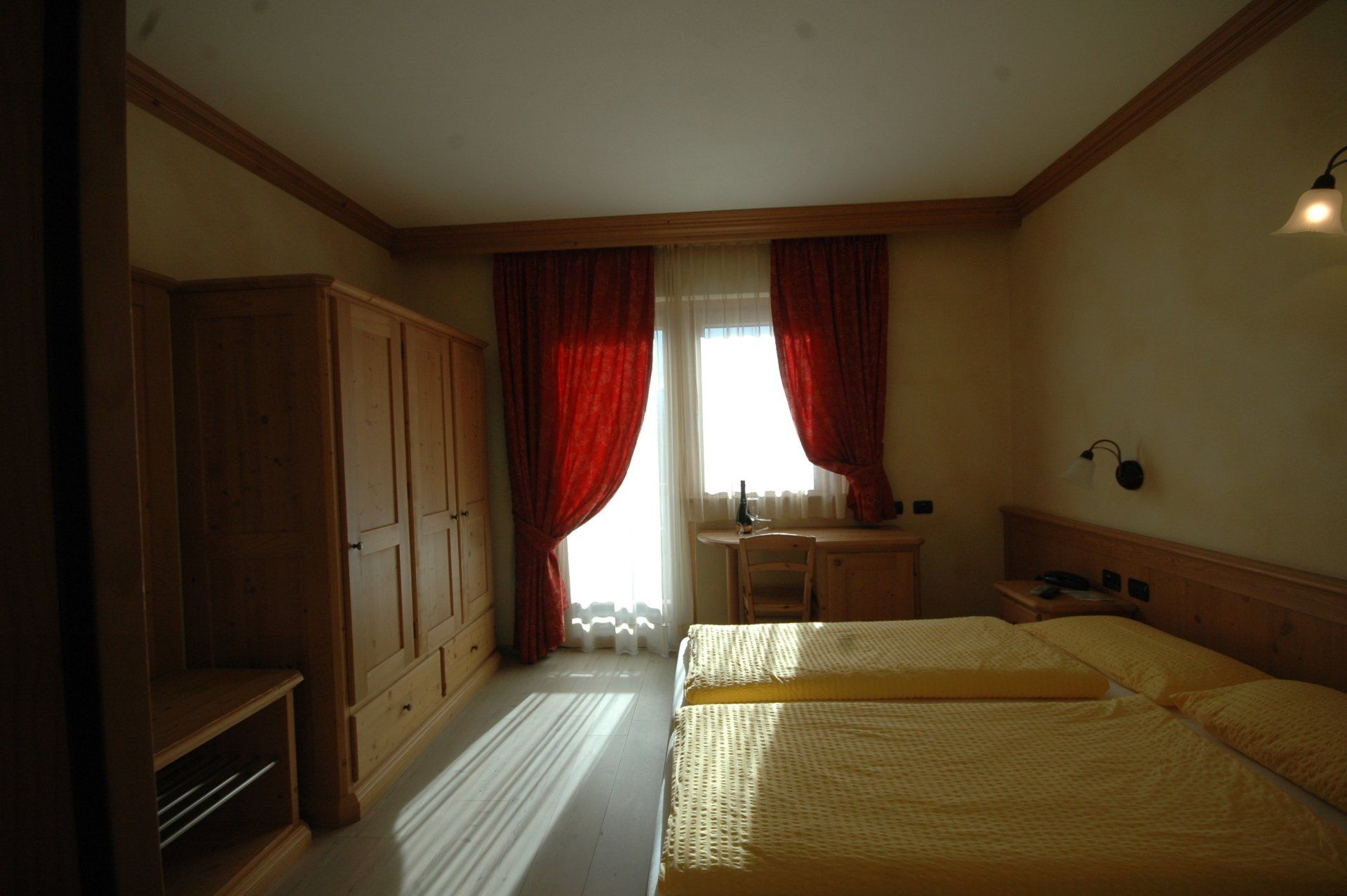 Camera da letto con due letti, mobili in legno, tende rosse e una finestra che lascia entrare la luce del sole.