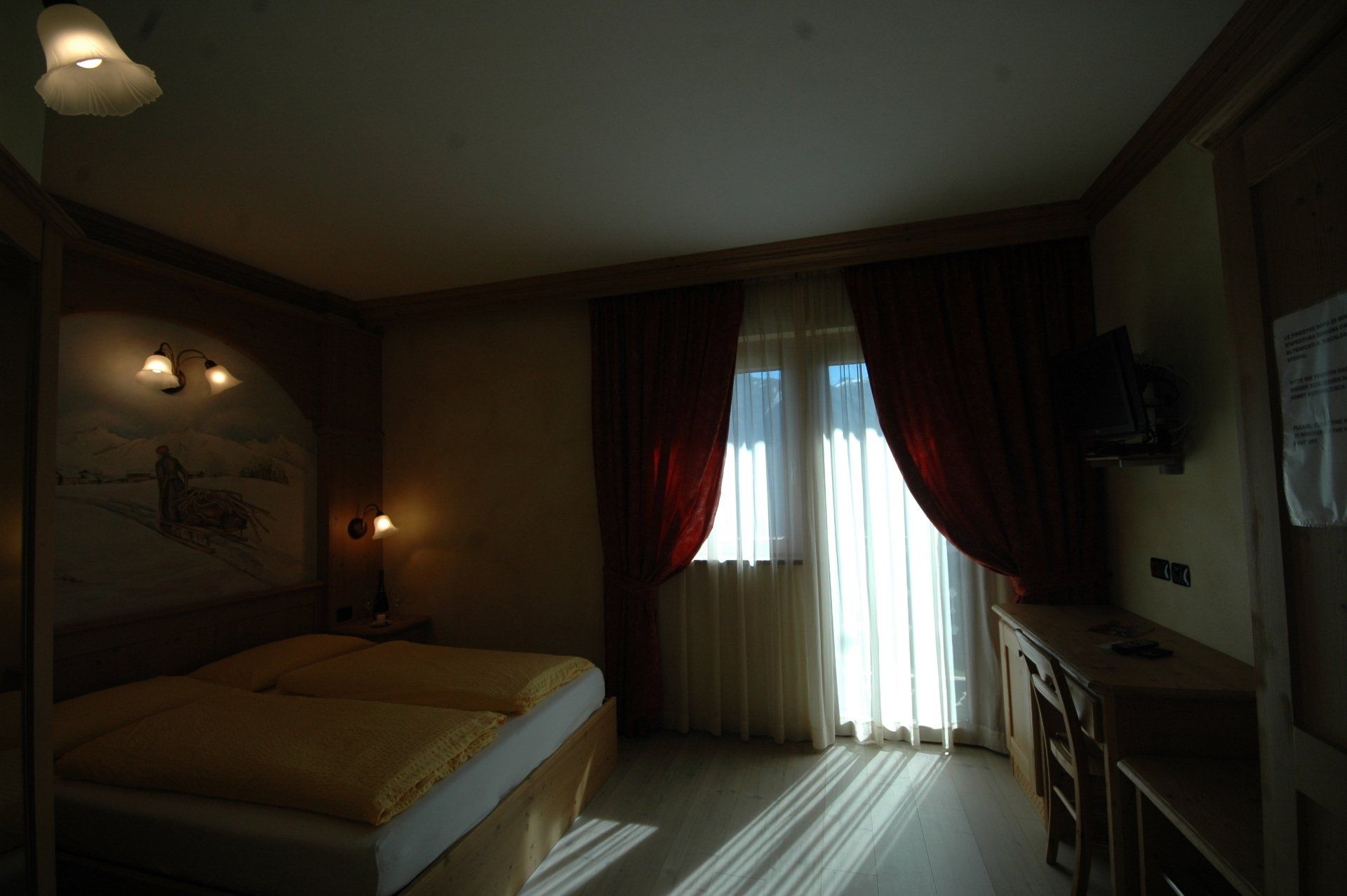 Interno della camera d'albergo: letto, finestra con tende, cassettiera, illuminazione calda.