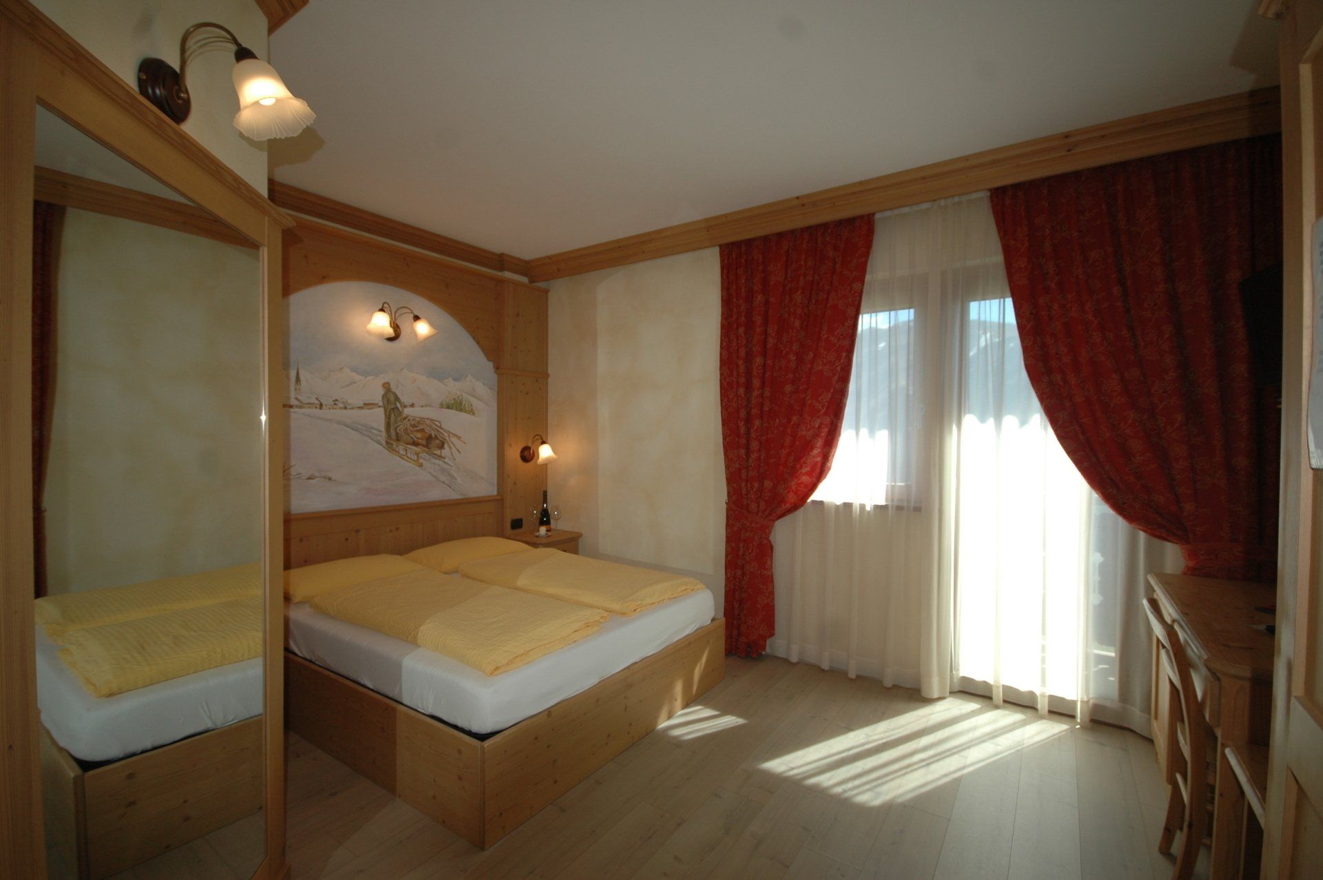 Camera da letto con letto, tende rosse, dettagli in legno e luce del sole che filtra dalla finestra.
