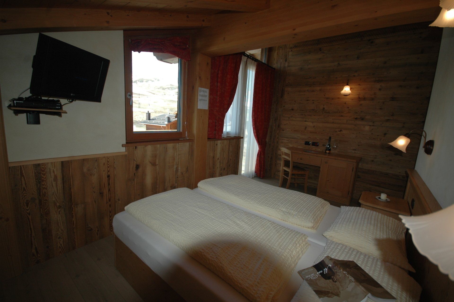 Accogliente camera da letto con due letti singoli, pareti in legno, piccola scrivania e TV. Finestra con tende rosse.
