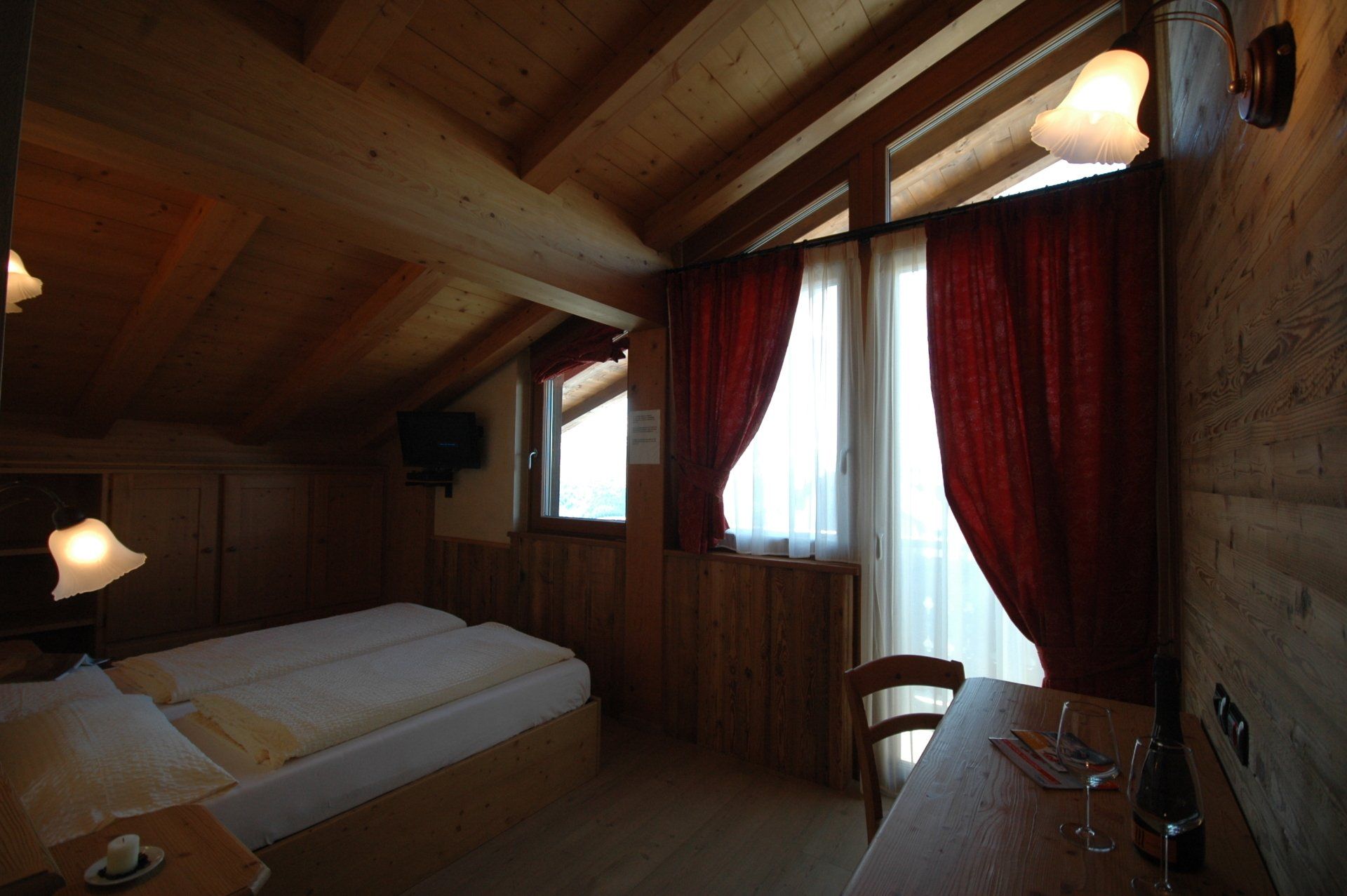 Camera da letto mansardata in legno con letto, tavolo, finestre e tende rosse.