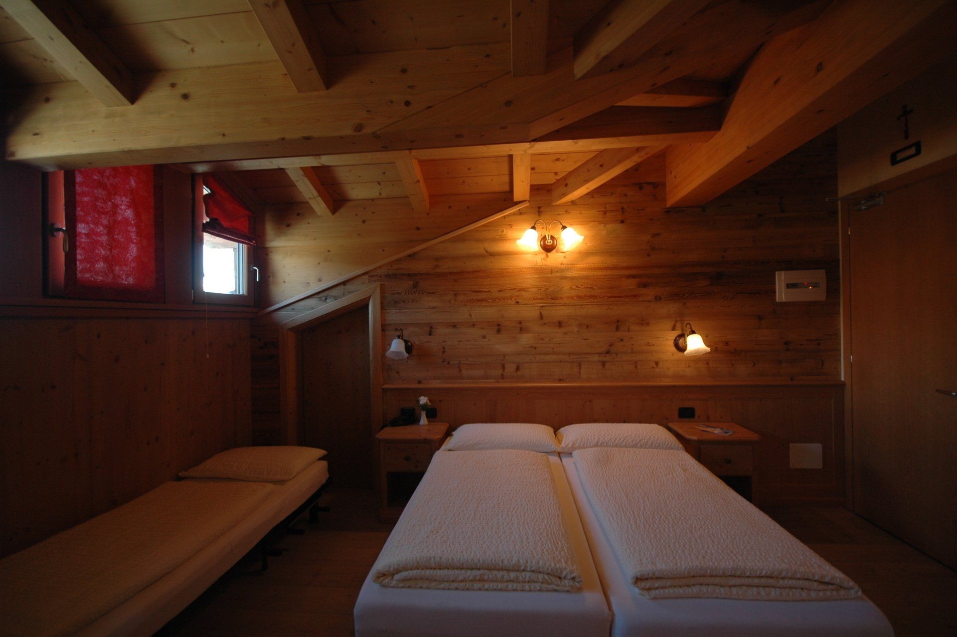 Camera da letto rustica con travi in ​​legno, un letto matrimoniale, un letto singolo e piccole finestre con tende rosse.