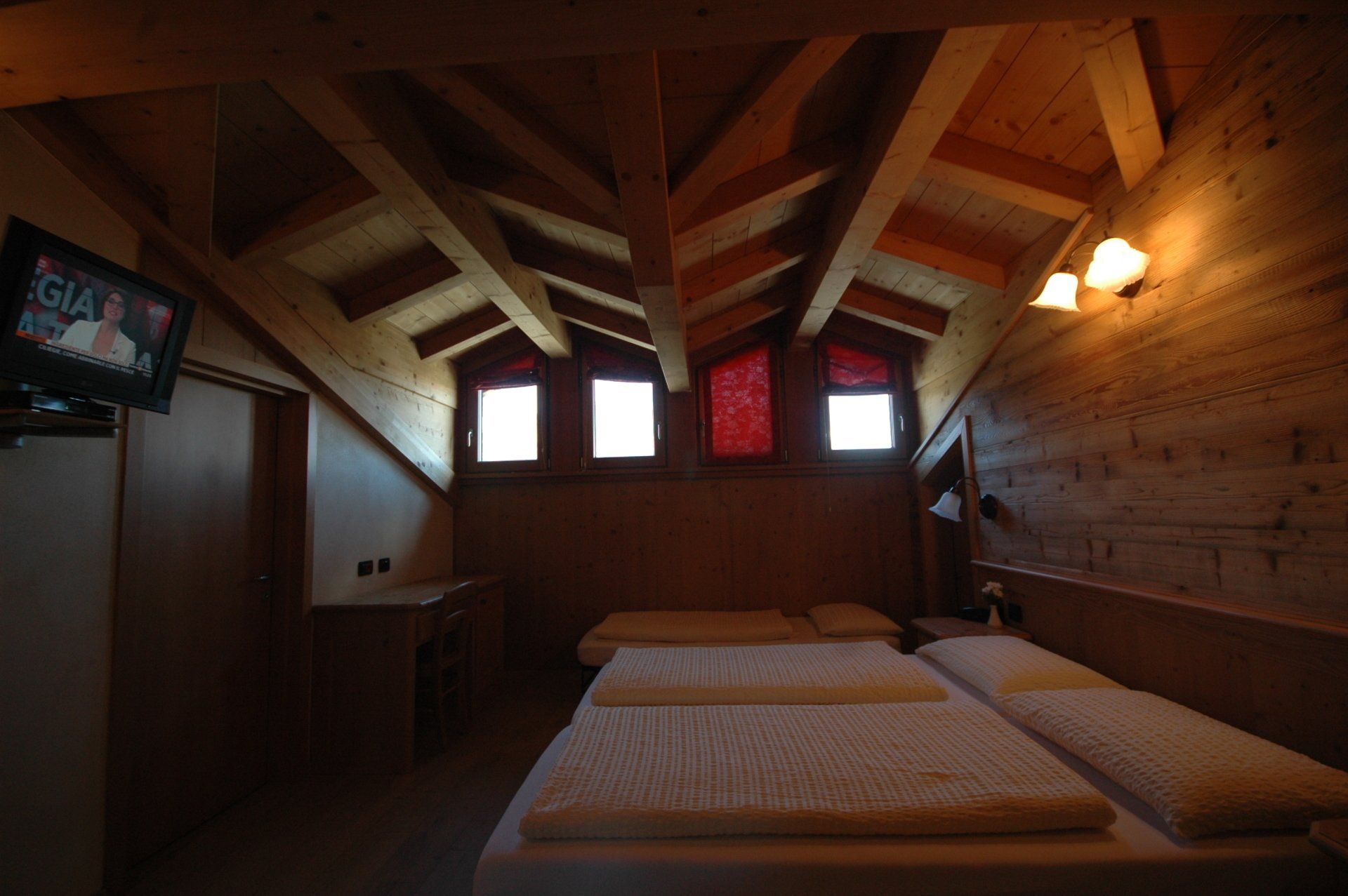 Camera da letto con soffitto in legno inclinato, piccole finestre, letto matrimoniale e TV.