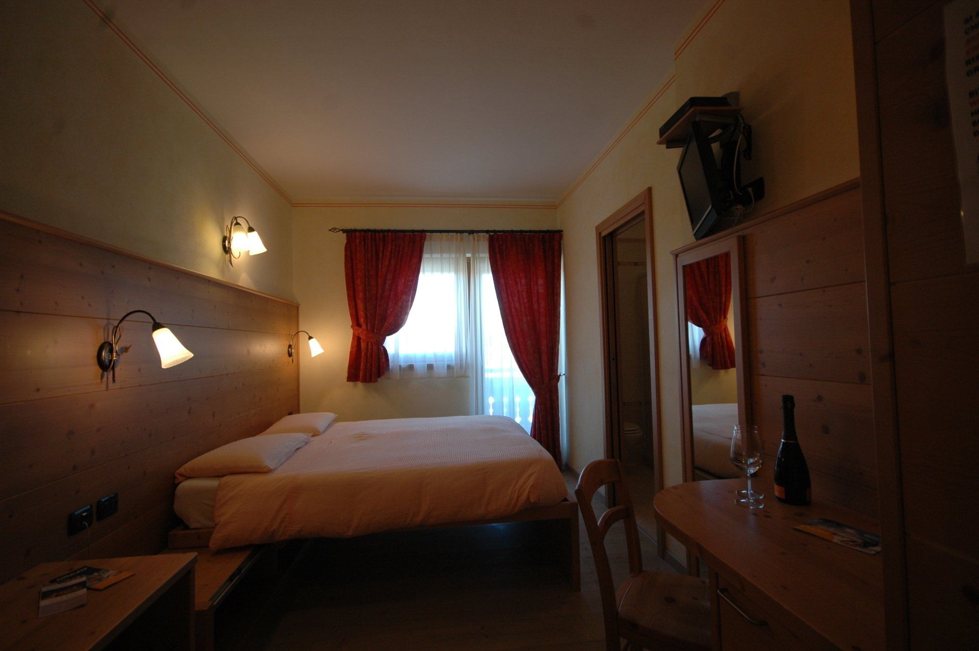 Camera d'albergo con letto, finestra con tende rosse, scrivania e televisione.