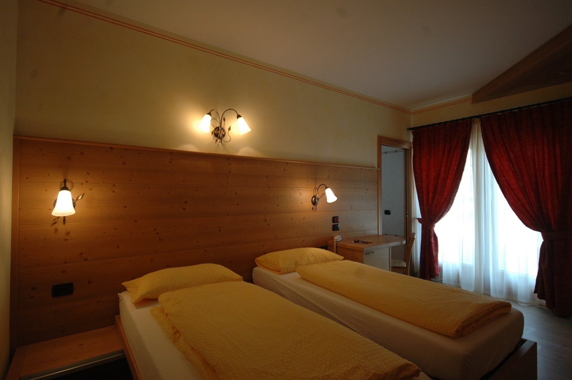Due letti singoli con lenzuola gialle in una stanza con testiera in legno, tende rosse e applique.