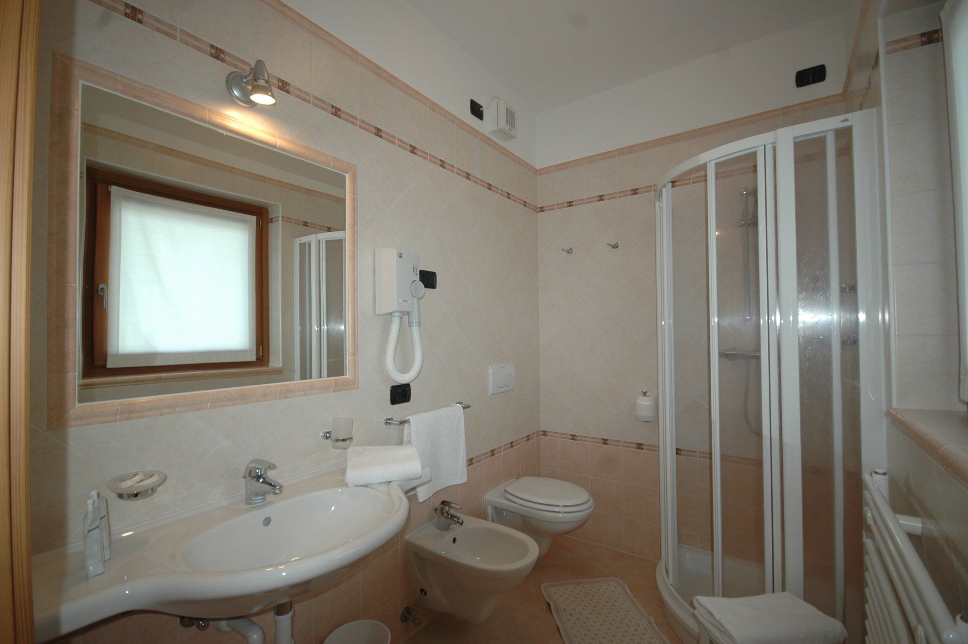 Bagno con lavandino, WC, bidet, doccia e finestra. Pareti beige, sanitari bianchi e specchio.