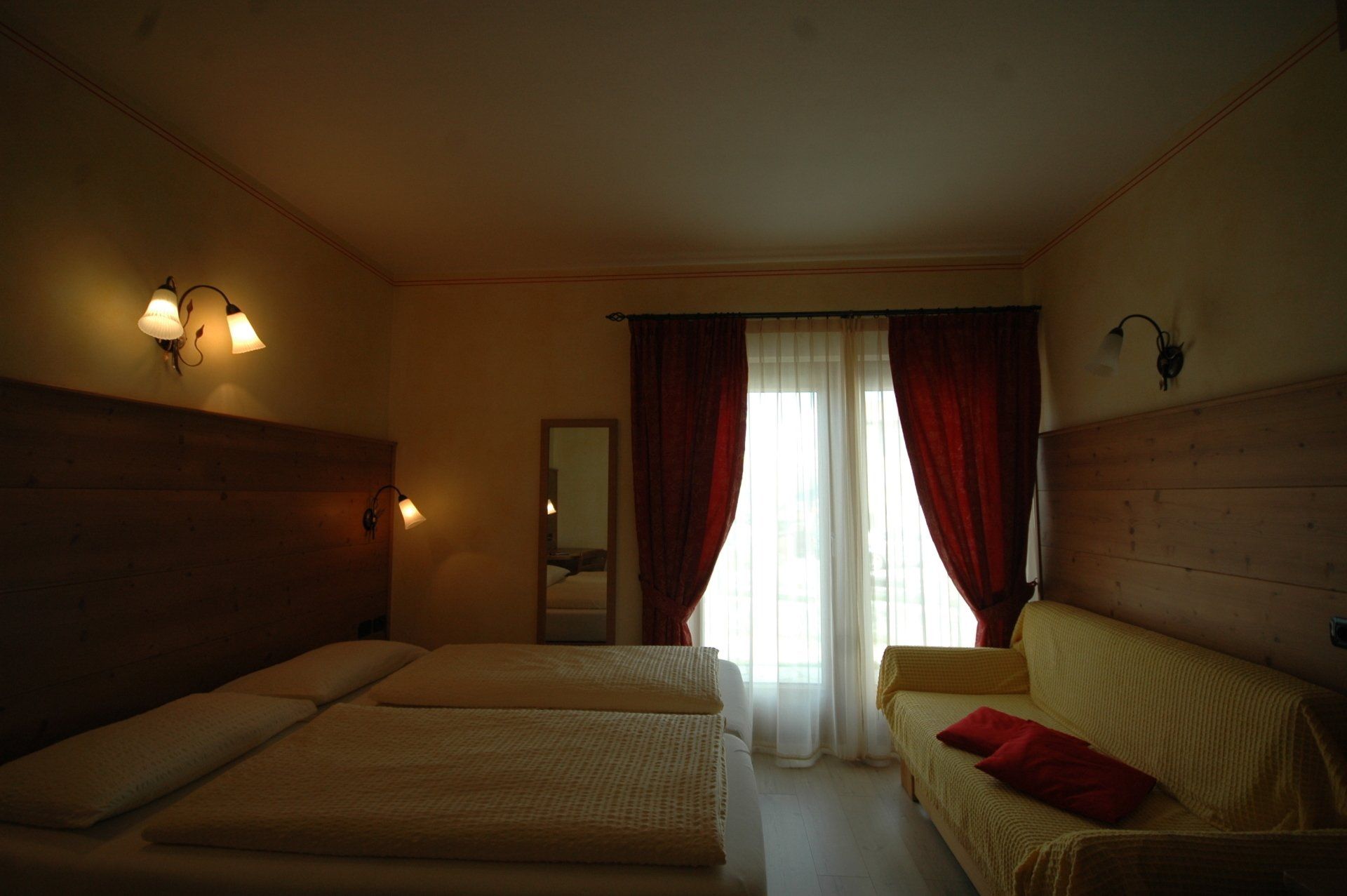 Camera da letto con letto matrimoniale, divano, finestra con tende rosse e specchio. Pareti beige e luci calde.