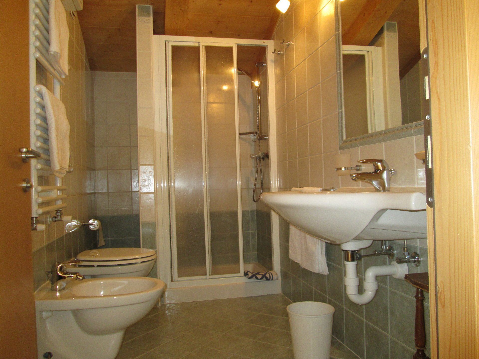 Bagno con WC, bidet, doccia, lavandino e portasciugamani. Piastrelle verde chiaro, soffitto in legno e porta doccia pieghevole.
