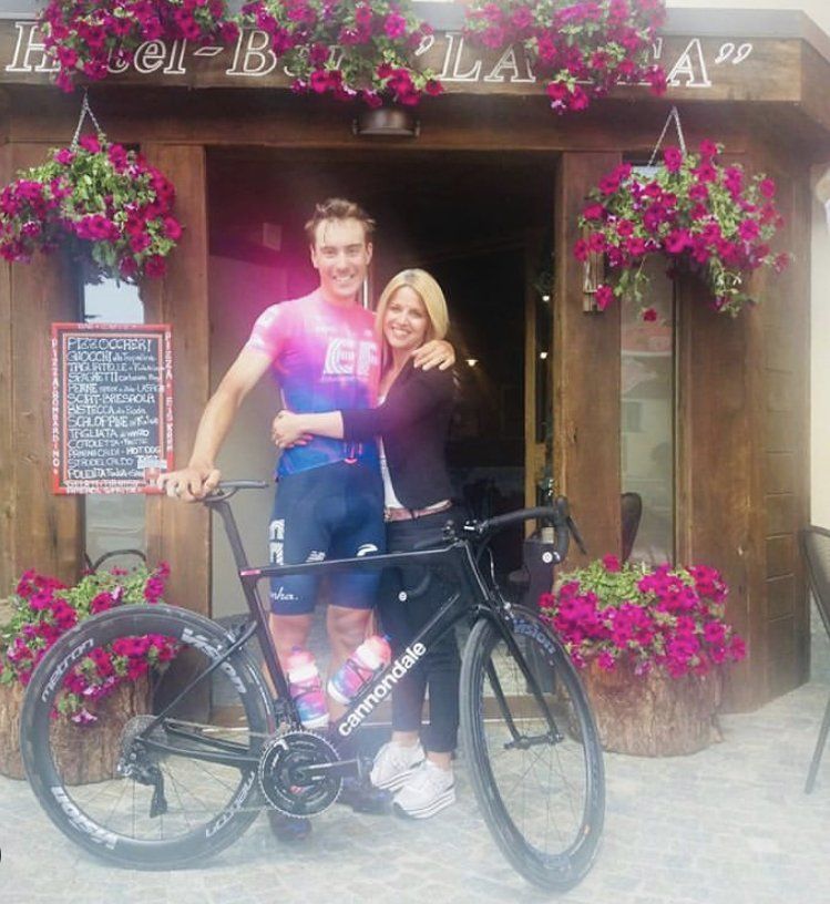 Ciclista in maglia rosa e blu con donna, in bicicletta davanti a un bar decorato con fiori.