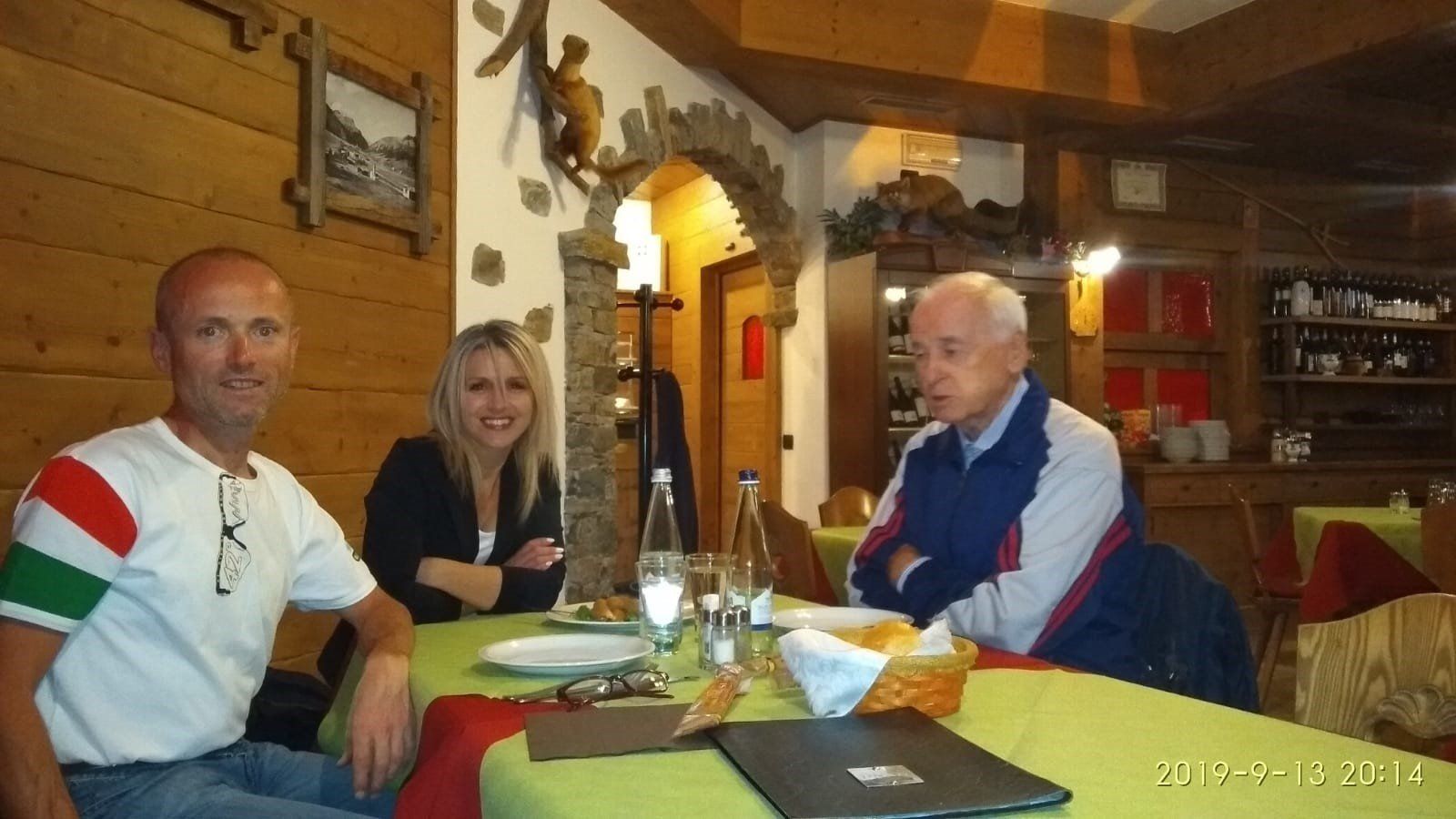 Tre persone sedute a un tavolo in un ristorante rustico. Un uomo in camicia bianca, una donna in blazer nero, un uomo anziano in giacca blu.