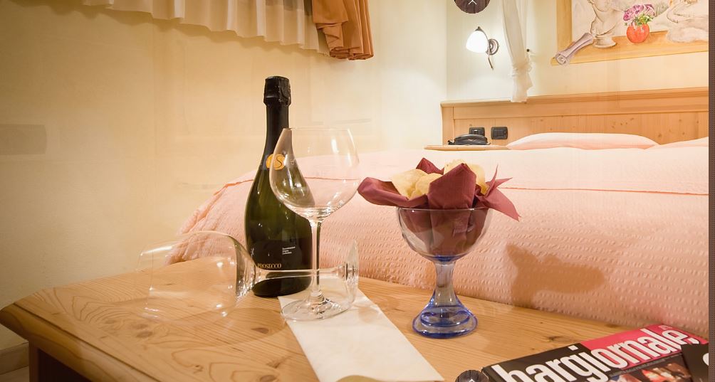 Bottiglia di champagne, bicchieri, fiori e una barretta di cioccolato su un comodino di legno davanti al letto.