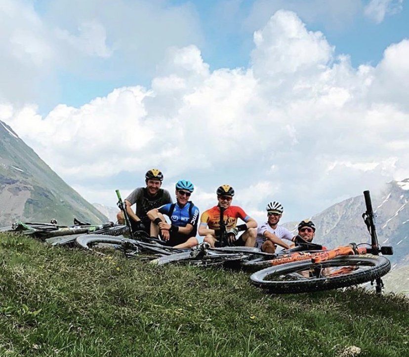 Mountain biker su una montagna erbosa, sorridenti, che si riposano con le loro bici, cielo nuvoloso, montagne innevate sullo sfondo.