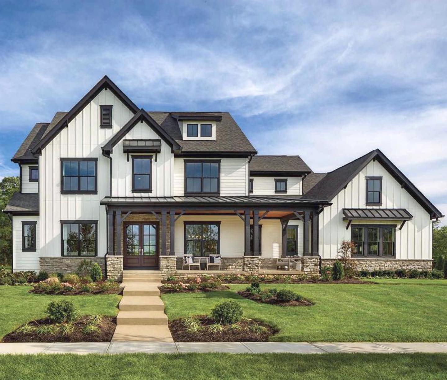 A two-story farmhouse with white vertical siding, dark window frames, a stone base, and a large front porch.