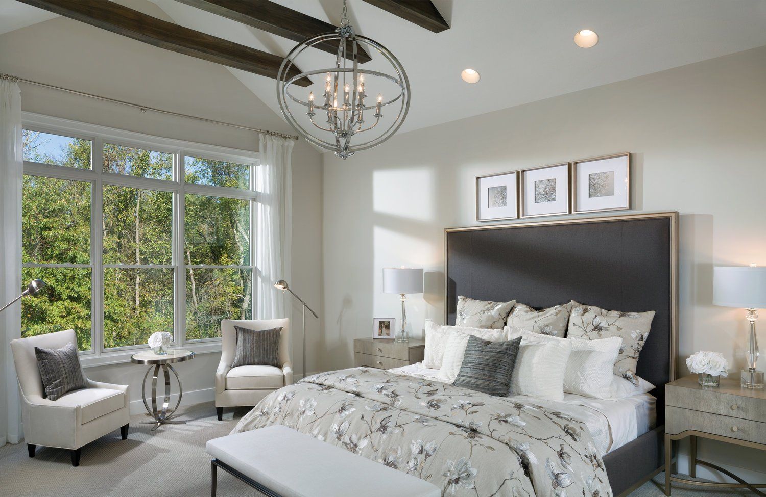 A modern, neutral-toned bedroom with a large gray upholstered headboard, floral bedding, chandelier, and two armchairs.