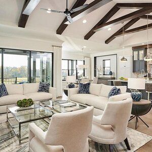 A bright, modern open-plan living room with white sofas, cream armchairs, a glass coffee table, and dark ceiling beams.