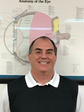 Man smiling, wearing a black vest and white collared shirt, stands in front of an anatomy of the eye chart.