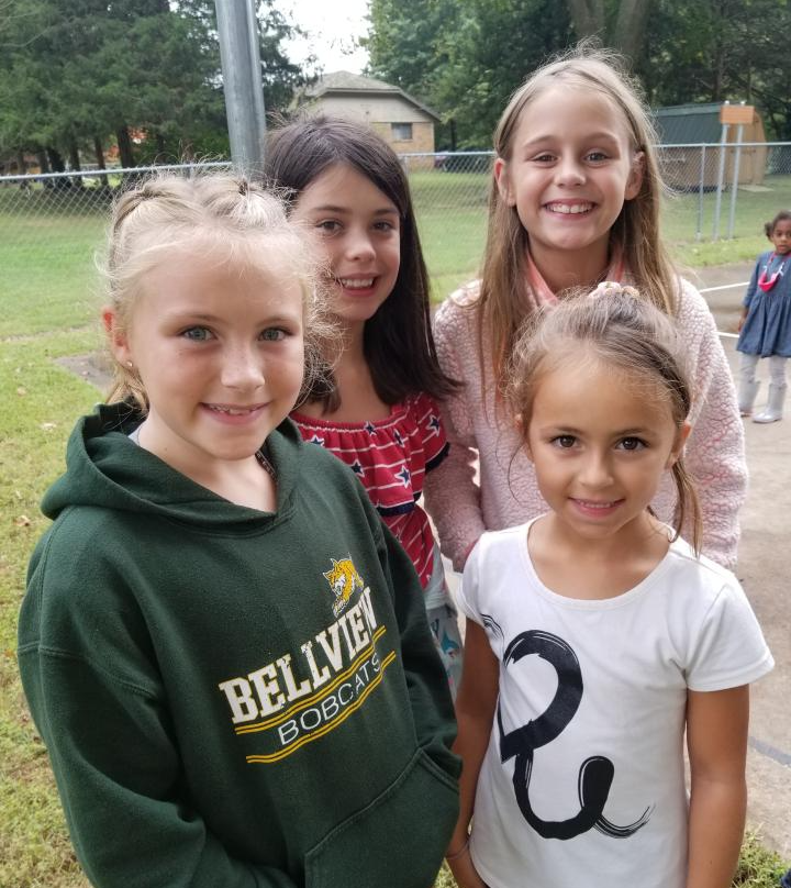 Four smiling girls in a park setting. One wears a green sweatshirt, another a white shirt with a design.