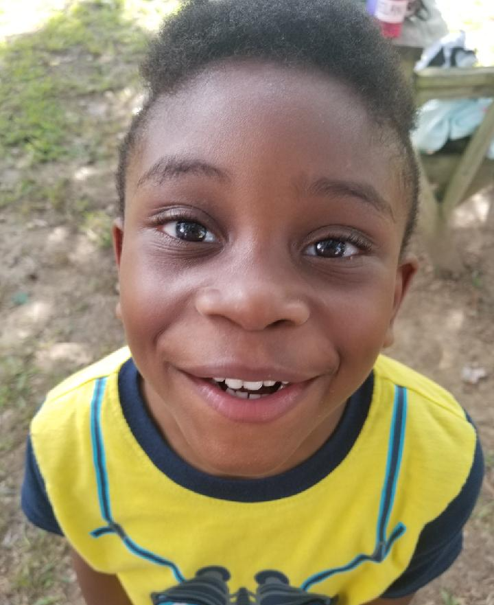 Smiling Black boy with dark skin, wearing yellow and blue shirt, looking at camera outside.