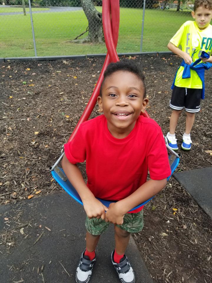 Boy in red shirt smiles, sitting in a playground swing. Another boy in yellow shirt stands nearby.