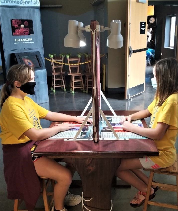 Two girls playing a game at a table with lights, in a science lab. One girl wears a mask.