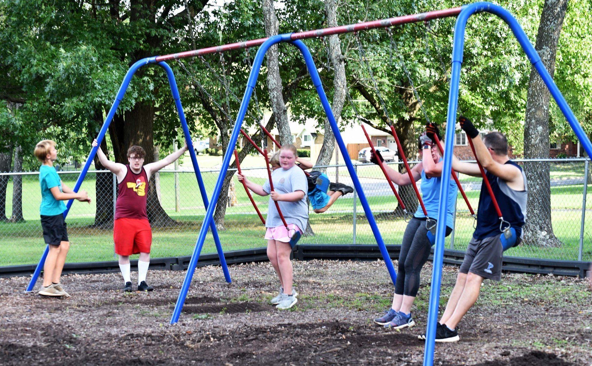 Children swing on a blue swing set at a park. Some are standing or smiling.