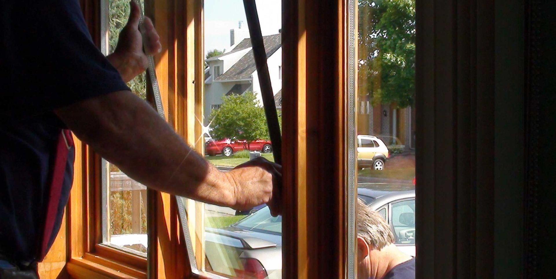 Un homme ouvre une fenêtre en bois à l'intérieur d'une maison, des voitures et des arbres sont visibles à l'extérieur.