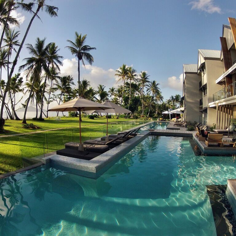 Poolside view of a resort with palm trees, sun loungers, and blue water under a sunny sky.