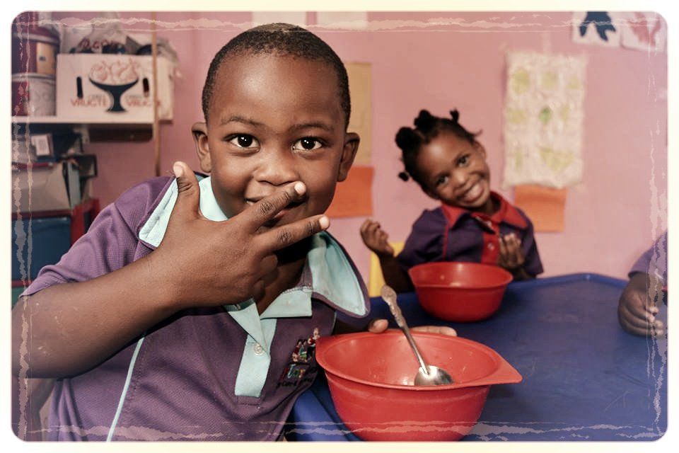 Two children eat from red bowls at a table, with one child smiling in the background.