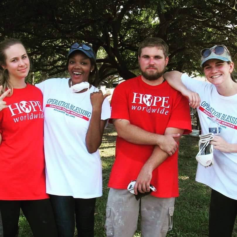 Four volunteers in red and white “HOPE worldwide” shirts posing outdoors under a tree
