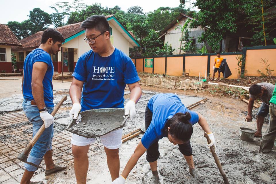 Volunteers in blue shirts and gloves mixing concrete at a muddy outdoor construction site