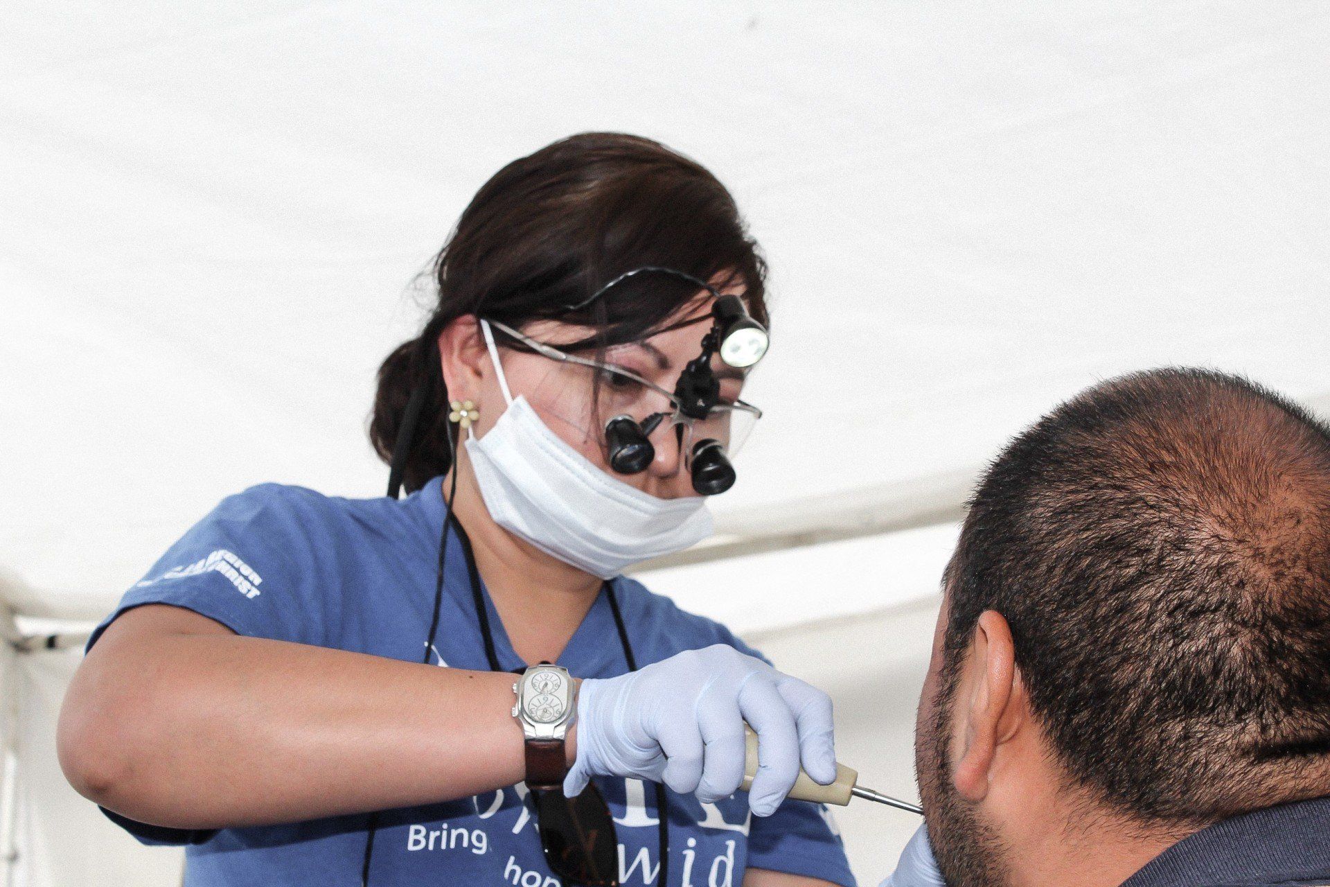 Dentist in blue scrubs wearing a face shield and mask treats a patient in a clinic