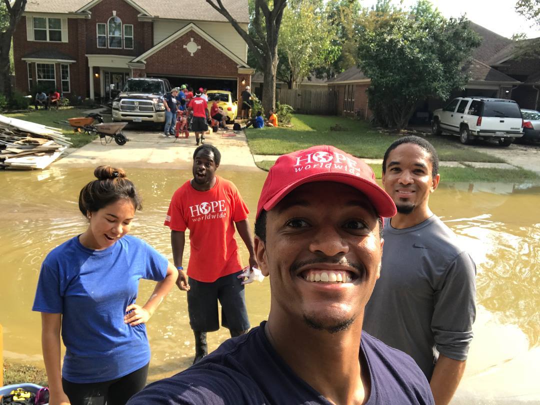 Four people standing in floodwater on a suburban street, smiling with damaged homes and vehicles behind them.