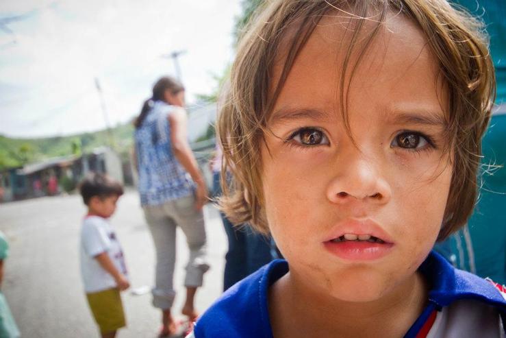 Close-up of a child in a blue shirt outdoors, with two blurred people and a building in the background