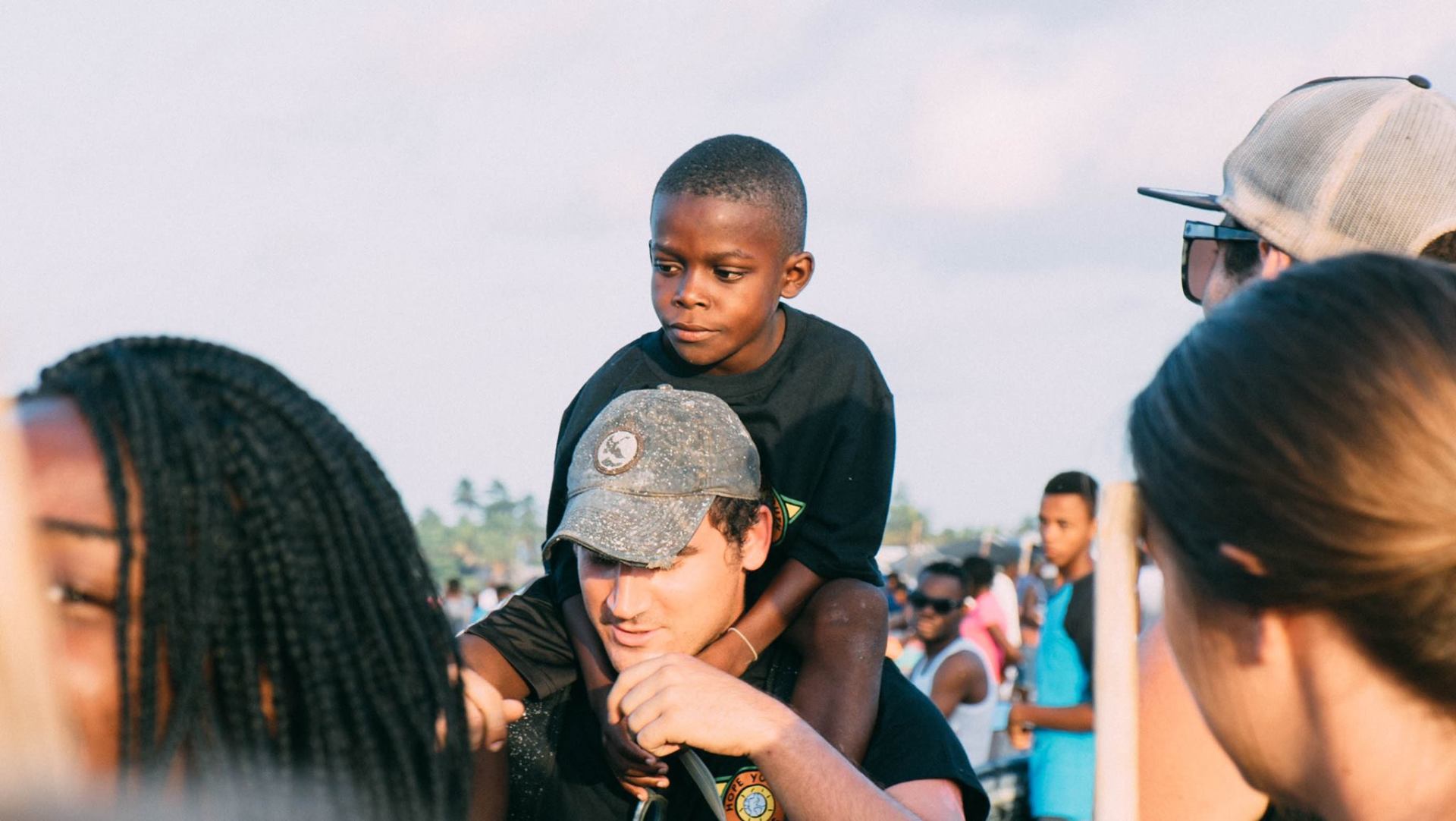 Child riding on an adult’s shoulders in a crowd outdoors at sunset