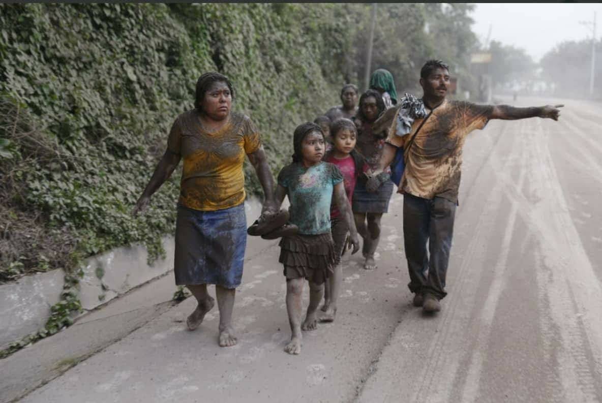 People walking along a wet roadside in heavy rain, carrying bags and pointing ahead.
