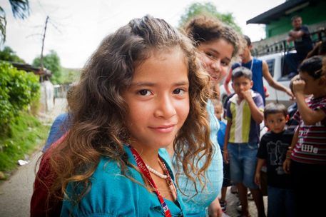 Smiling child in a turquoise shirt outdoors, with other children and a rural house in the background